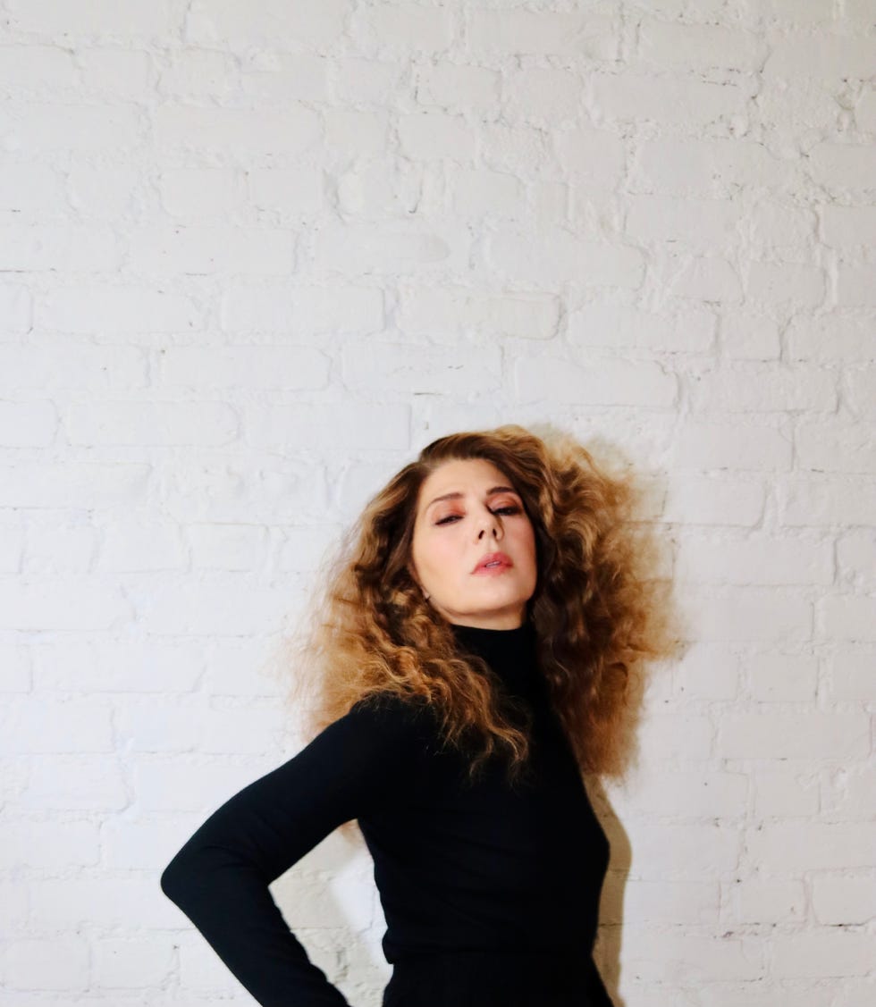 person with curly hair posing against a white brick wall