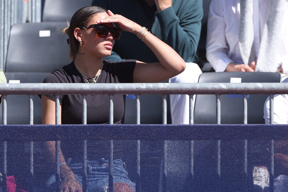 a woman sits in stands behind a metal fence and holds her hand over her eyes as she looks right