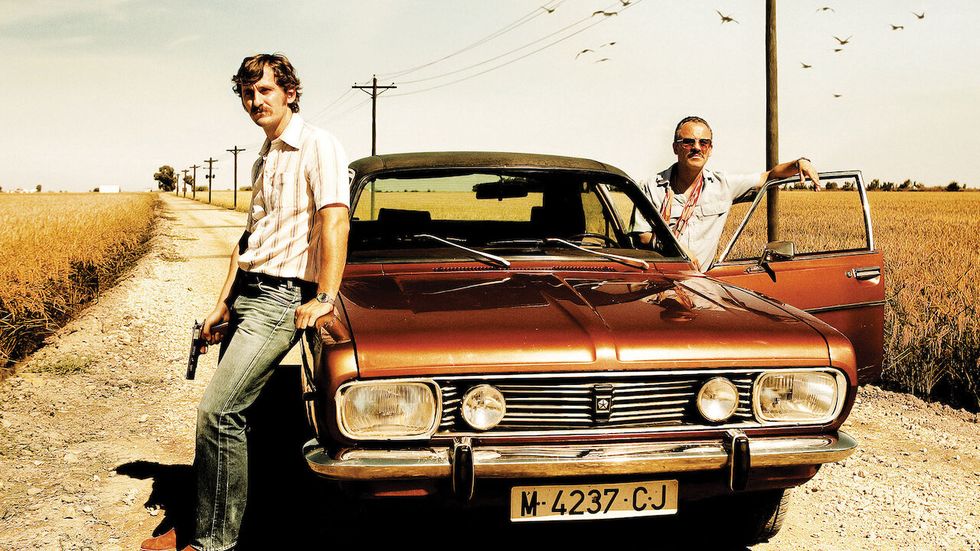 a couple of men posing by a car on a dirt road