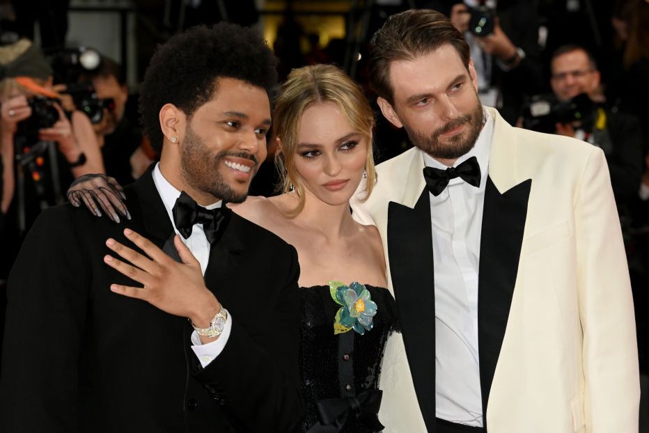the weeknd, wearing a black tuxedo, lily rose depp, wearing a green dress, and sam levinson, wearing a white tuxedo, posing for a photo together as photographers take pictures behind them