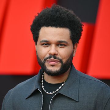 a man with a small afro and beard smiles at the camera, he wears a gray zip up jacket black shirt and silver chain necklace