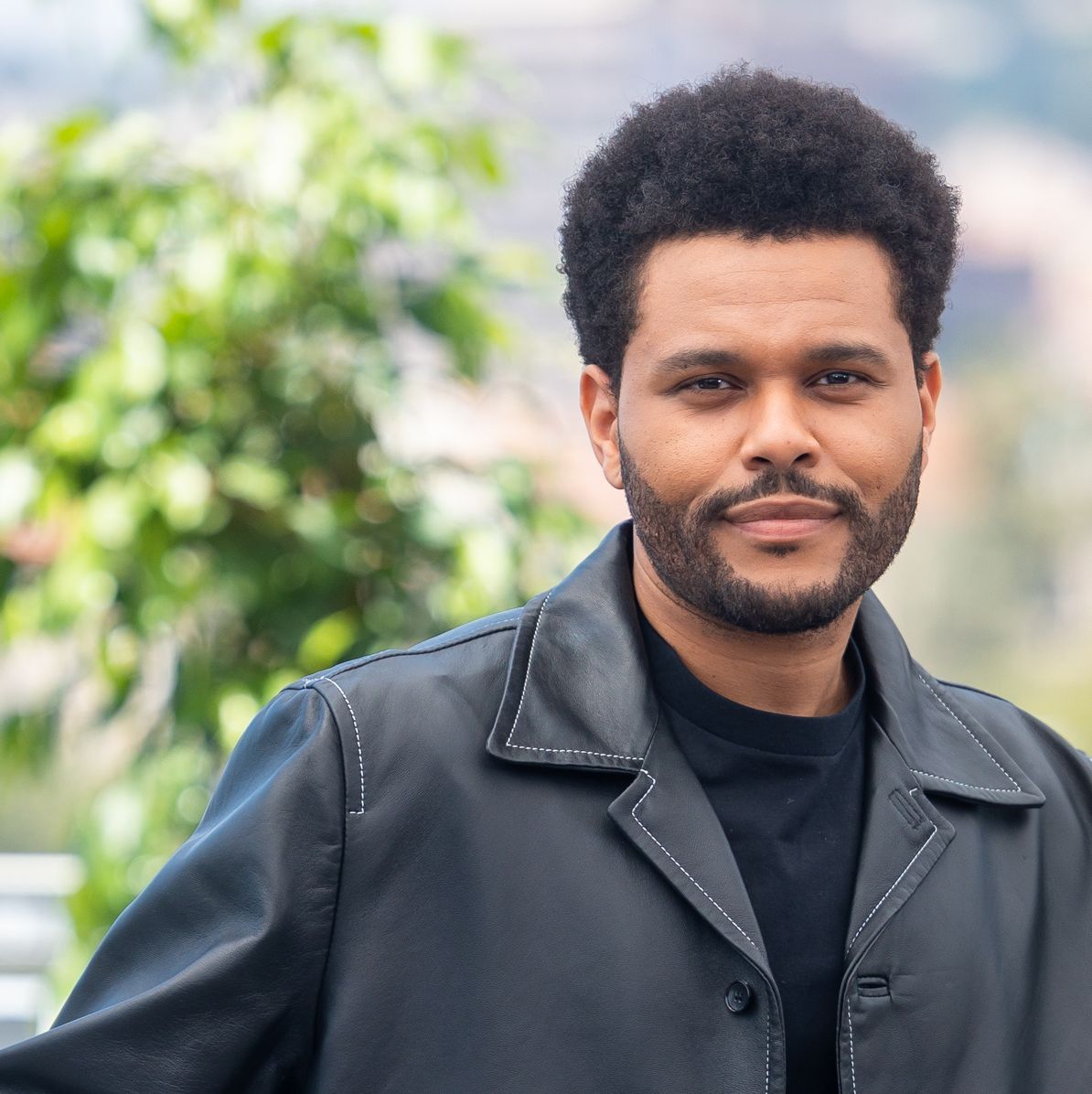 the weeknd smiles at the camera while standing outside in a black jacket and black shirt