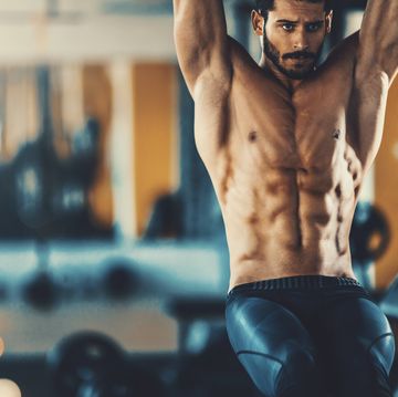 closeup front view of a handsome mid 20's man doing abs in a gym he's doing leg raises, shirtless blurry gym equipment in background