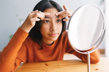 asian young woman squeezing acne skin and looking on the mirror at home, close up of young women face