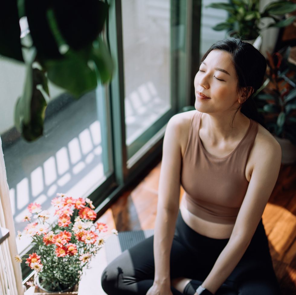 active young asian sports woman taking a break after working out at home, sitting on exercise mat taking a deep breath with her eyes closed. sports and exercise routine. health, fitness and wellness concept