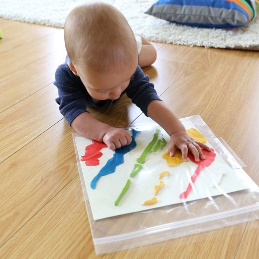 a toddler doing tummy time squishes finger paint that's inside a plastic freezer bag