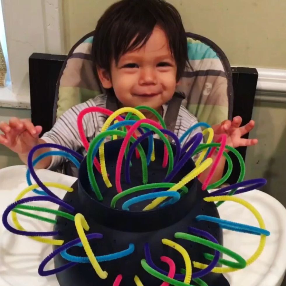 a toddler looks at a colander that has pipe cleaners laced through the holes