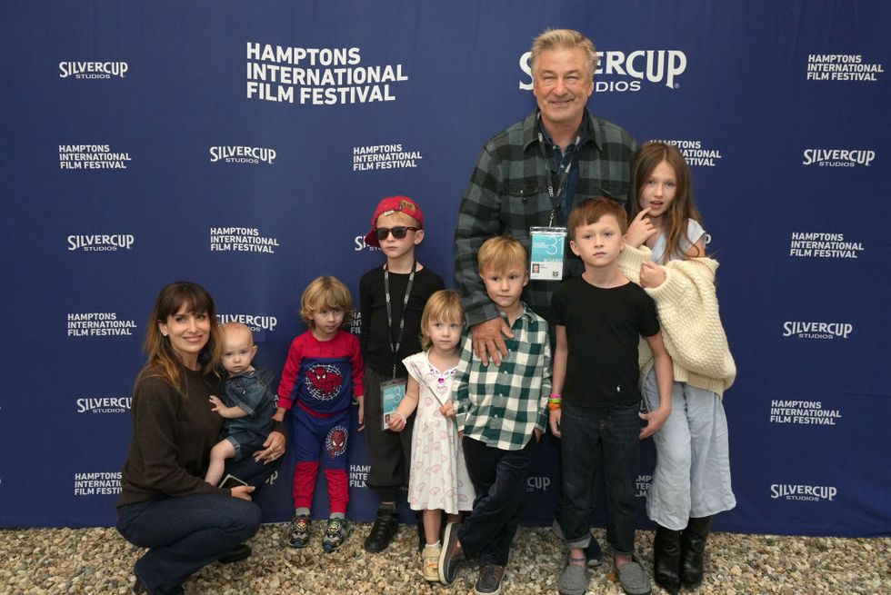 alec baldwin and his family pose for a photo in front of a blue background, his wife hilaria crouches on the left while holding their youngest child, alec smiles with standing among six other children