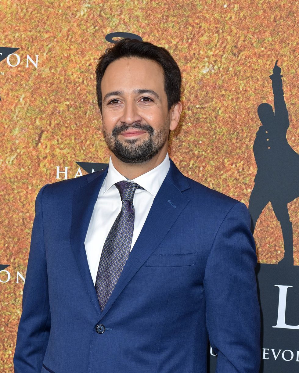 a man in a blue suit and gray tie smiles while standing in front of a golden background with repeating star icons