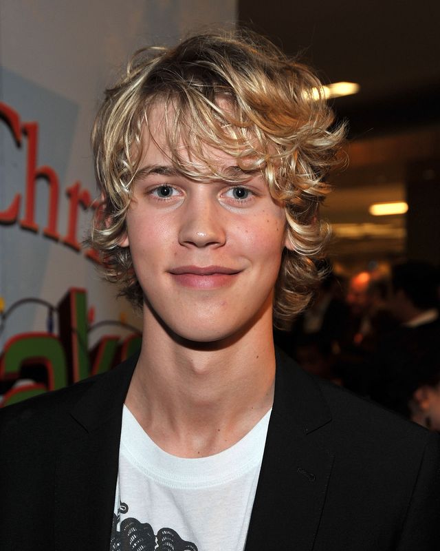 austin butler smiles at the camera, he wears a black cardigan over a white graphic tshirt