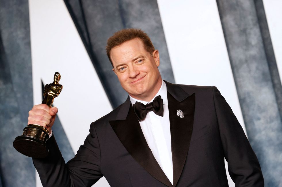 brendan fraser, wearing a black tuxedo, smiles and holds an academy award in one hand, standing in front of a blue and white backdrop