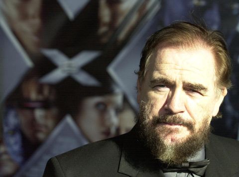 brian cox, wearing a black tuxedo and with a beard, stands in front of a poster for x2 x men united