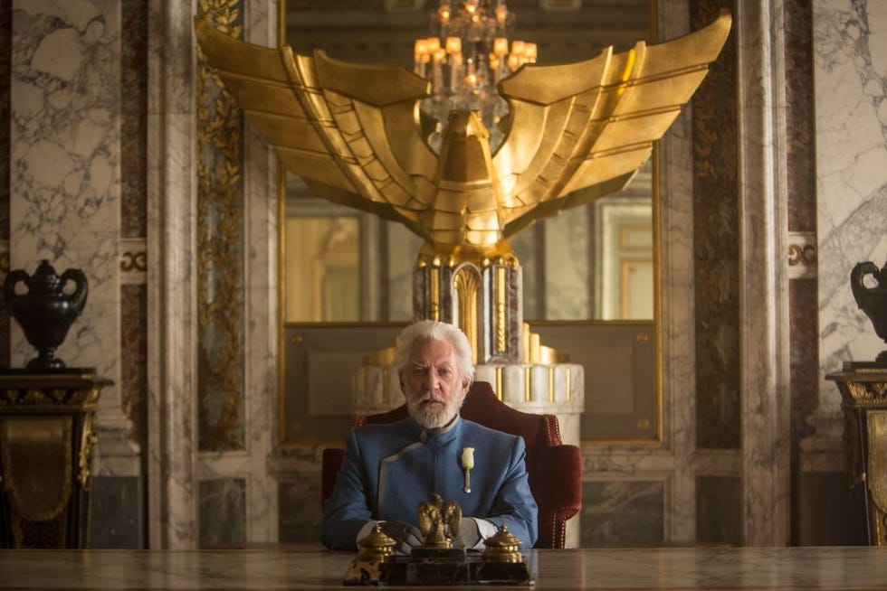 donald sutherland sits in costume for the hunger games mockingjay part one, he wears a blue suit with a rose attached to his brest