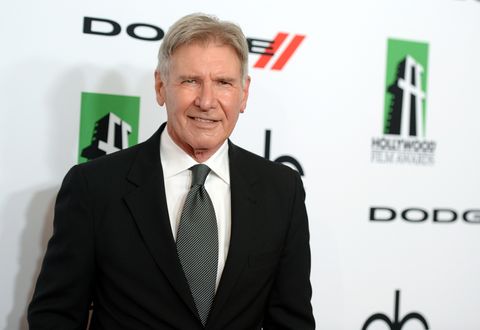 harrison ford, wearing a black suit jacket, white shirt, and gray tie, smiles while standing in front of a white wall with several logos on it
