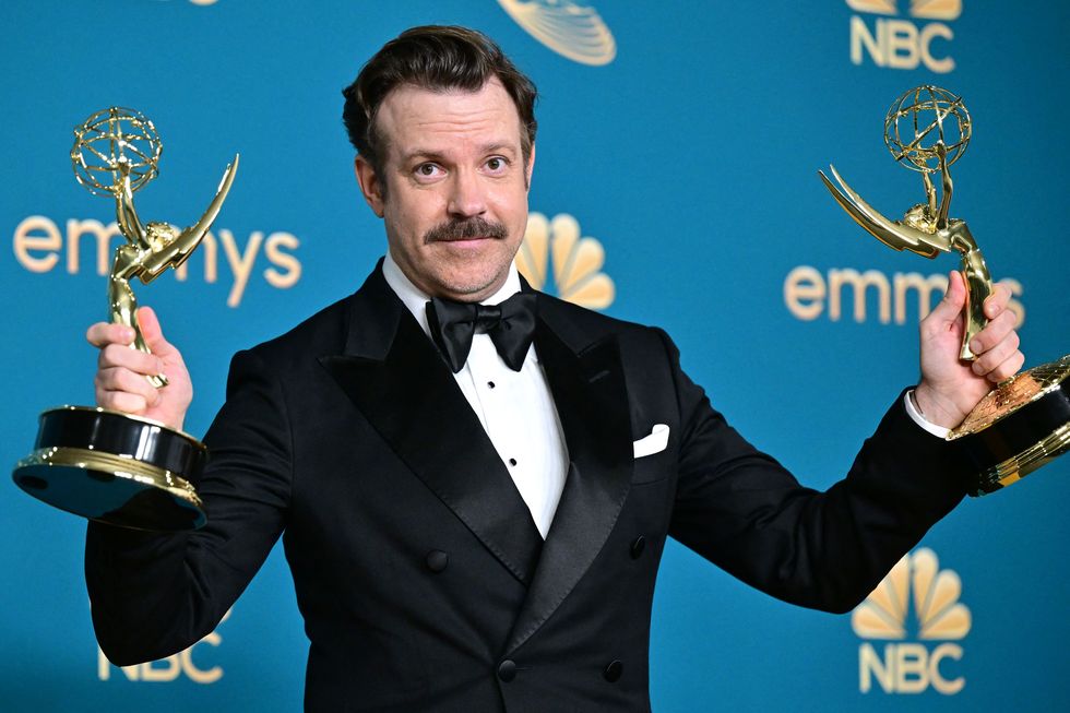 jason sudeikis, wearing a black tuxedo, holds an emmy award in each hand as he stands in front of a blue backdrop