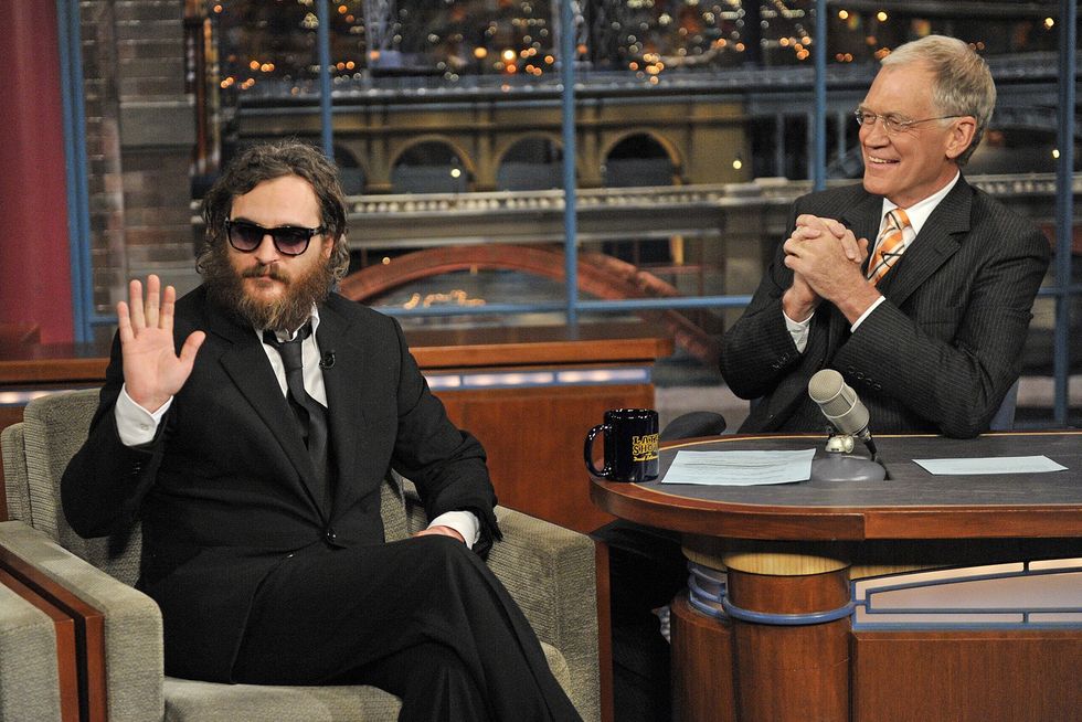joaquin phoenix waves while sitting in an armchair on a tv set and wearing a black suit and black sunglasses, to the right sits david letterman behind a desk with his hands clasped, letterman looks at phoenix and smiles