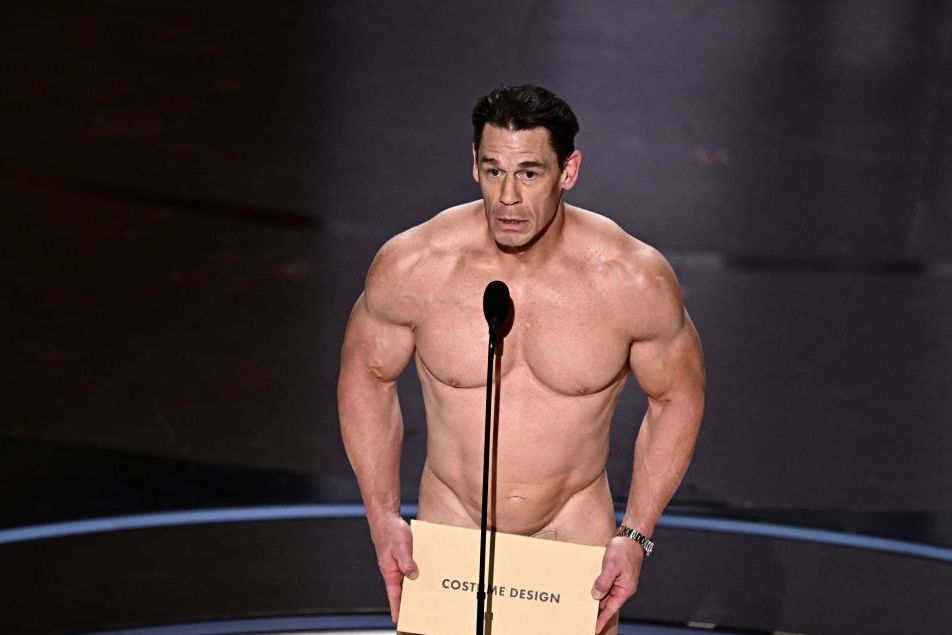 john cena stands at a microphone without clothes and holds a envelope that says costume design in front of his groin