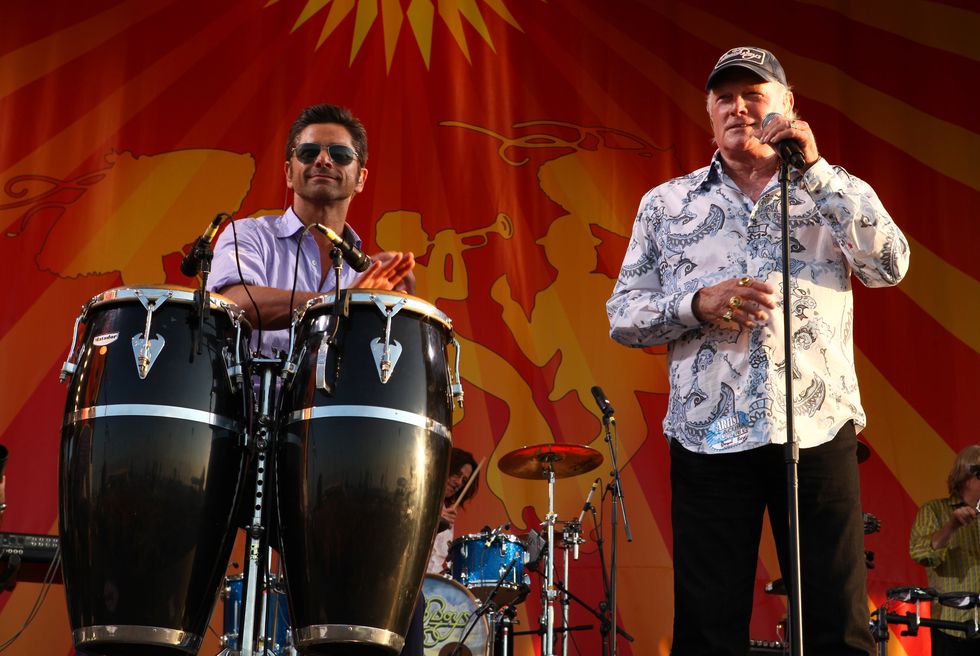 john stamos plays percussion on stage while wearing sunglasses and a purple short sleeved collared shirt, mike love of the beach boys stands to the right, love holds a microphone on a stand in one hand and wears black pants, a white paisley patterned shirt and a baseball cap