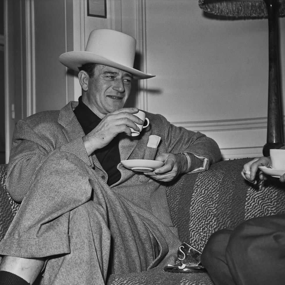actor john wayne sits on a sofa drinking a hot drink from a cup and saucer, while wearing a cowboy hat, february 26th 1951. (photo by powell/daily express/hulton archive/getty images)