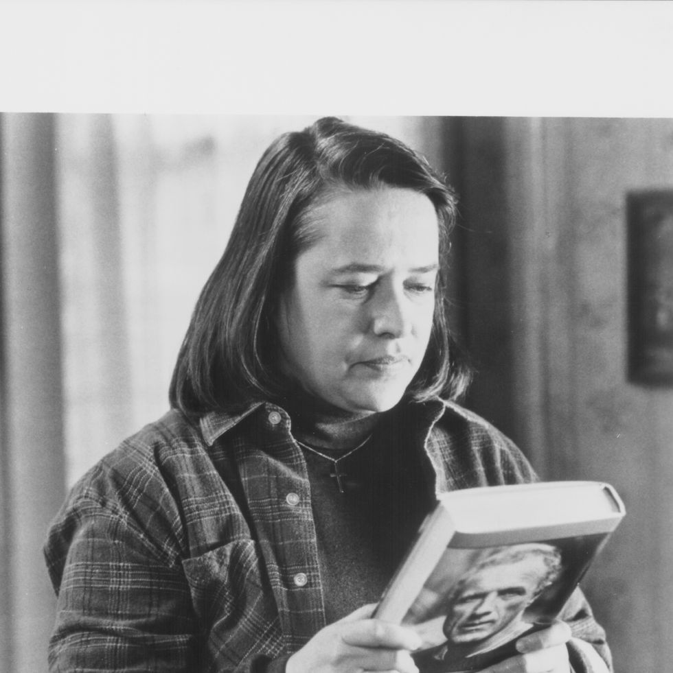 a woman looks down at a book she holds in her hands, she wears a plaid shirt over a sweater with a cross necklace