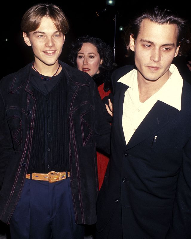 leonardo dicaprio and johnny depp stand next to each other, dicaprio smiles and wears a denim jacket, black vest, black shirt and royal blue pants with a brown belt, depp leans toward the camera and wears a black suit with a white collared shirt