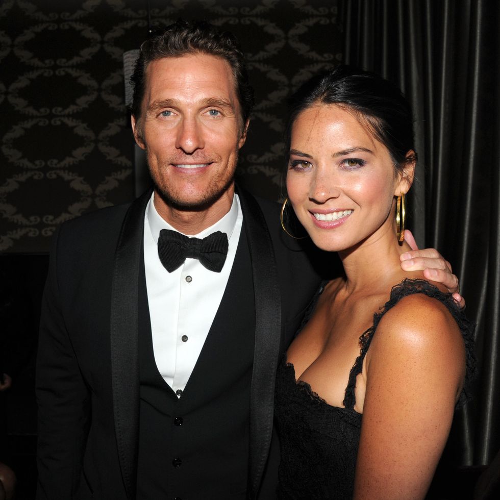 matthew mcconaughey and olivia munn﻿ smile for a photo while standing together in a room, he wears a black tuxedo, she wears a sleeveless black dress and hoop earrings