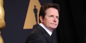 michael j fox stands on the red carpet at the oscars, he is wearing a black suit and tie with a white collared shirt and stands in front of a backdrop with an oscars logo on it, he looks past the camera with a neutral expression on his face