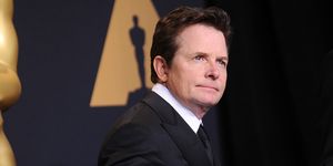 michael j fox stands on the red carpet at the oscars, he is wearing a black suit and tie with a white collared shirt and stands in front of a backdrop with an oscars logo on it, he looks past the camera with a neutral expression on his face