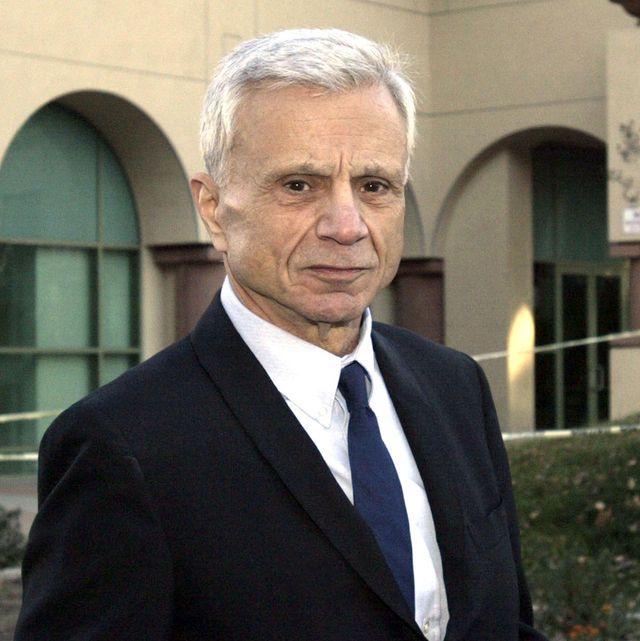 robert blake looks at the camera with a solemn expression, he is wearing a black suit coat, white collard shirt, and navy blue tie, he is outside standing in front of a tan building with curved arches over large windows