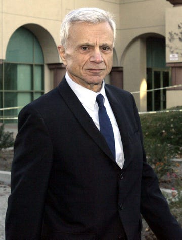 robert blake looks at the camera with a solemn expression, he is wearing a black suit coat, white collard shirt, and navy blue tie, he is outside standing in front of a tan building with curved arches over large windows