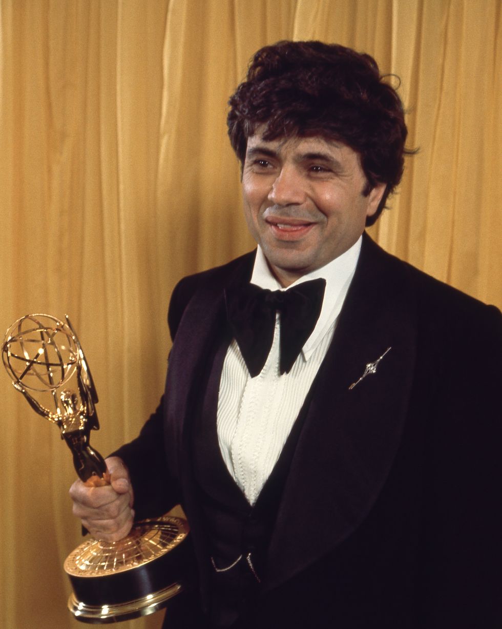 a 41 year old robert blake holds his emmy award in right hand, he is smiling and looking off camera while wearing a black tuxedo, white collared shirt, and large black bow tie, he is standing in front of a golden cloth backdrop