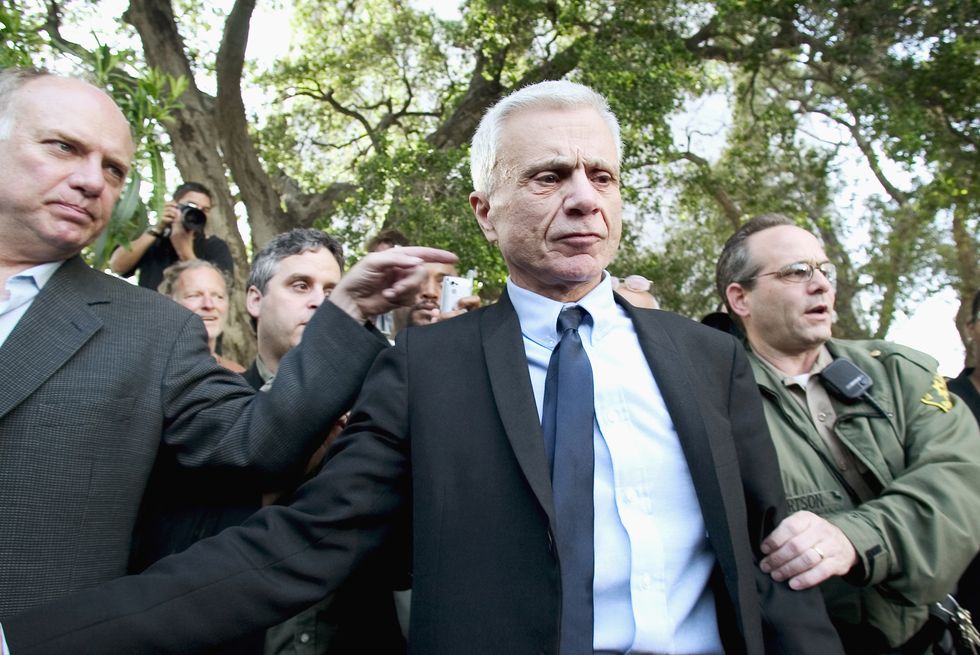 robert blake looks down and away from the camera while being surrounded by other people, he is wearing a black suit jacket, white collard shirt, and navy blue tie, to his right is a police officer, and a camera person is seen behind his left shoulder, tree canopies are also in the background
