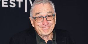 robert de niro stands at a clear podium with two microphones attached and smiles at the camera, he wears a green polo shirt, black suit jacket, and gray glasses