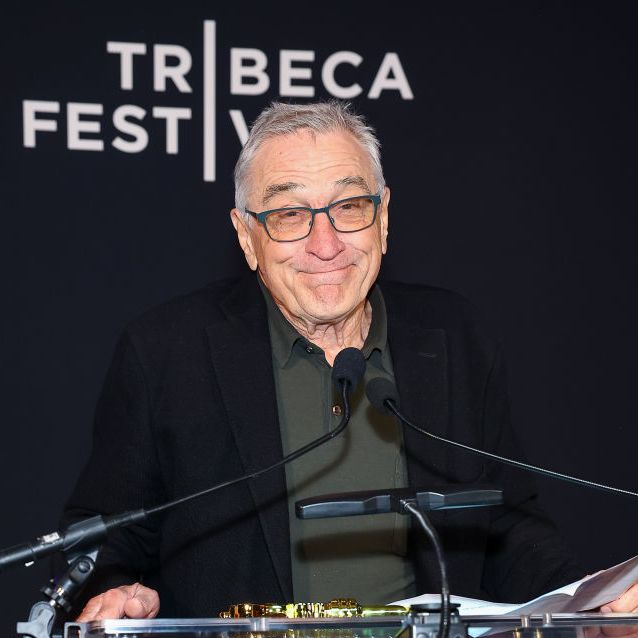 robert de niro stands at a clear podium with two microphones attached and smiles at the camera, he wears a green polo shirt, black suit jacket, and gray glasses