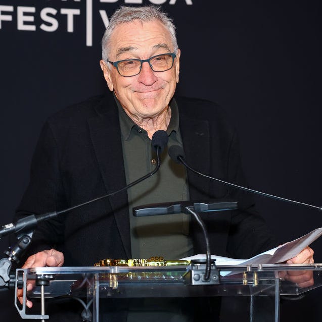 robert de niro stands at a clear podium with two microphones attached and smiles at the camera, he wears a green polo shirt, black suit jacket, and gray glasses