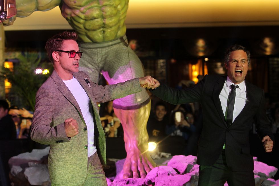 robert downey jr and mark ruffalo pose for photos while bumping fists in front of a green hulk statue, both men wear suits, ruffalo also wears a tie