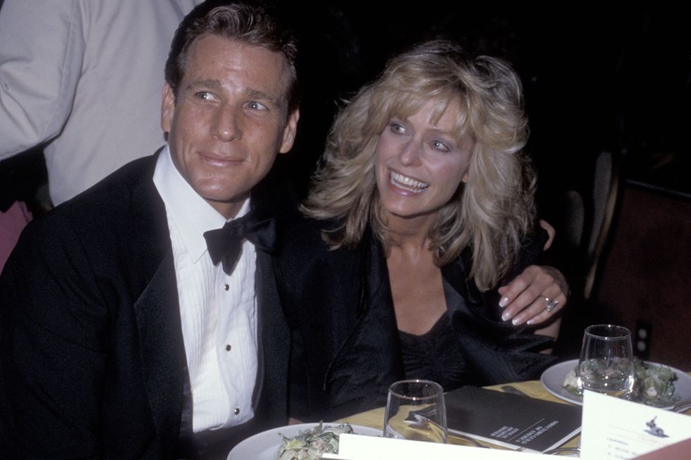 ryan o'neal and farah fawcett sit at a table and look to the left, he wears a black tuxedo, she wears a black outfit