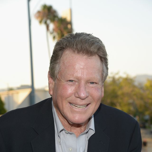 ryan o neal smiling for a photo, he wears a suit jacket and collared button up shirt and is outside with greenery in the background