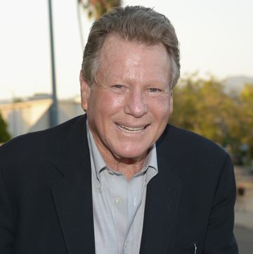 ryan o neal smiling for a photo, he wears a suit jacket and collared button up shirt and is outside with greenery in the background
