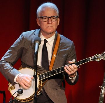 steve martin plays banjo on a stage in front of red curtain, he wears a gray suit with a black tie and white shirt, his glasses are black rimmed