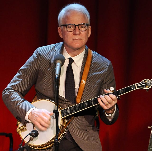 steve martin plays banjo on a stage in front of red curtain, he wears a gray suit with a black tie and white shirt, his glasses are black rimmed