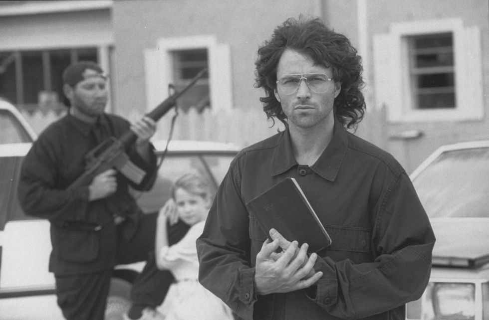 actor tim daly portraying david koresh in a publicity still for a movie, wearing a black shirt and glasses, holding a bible