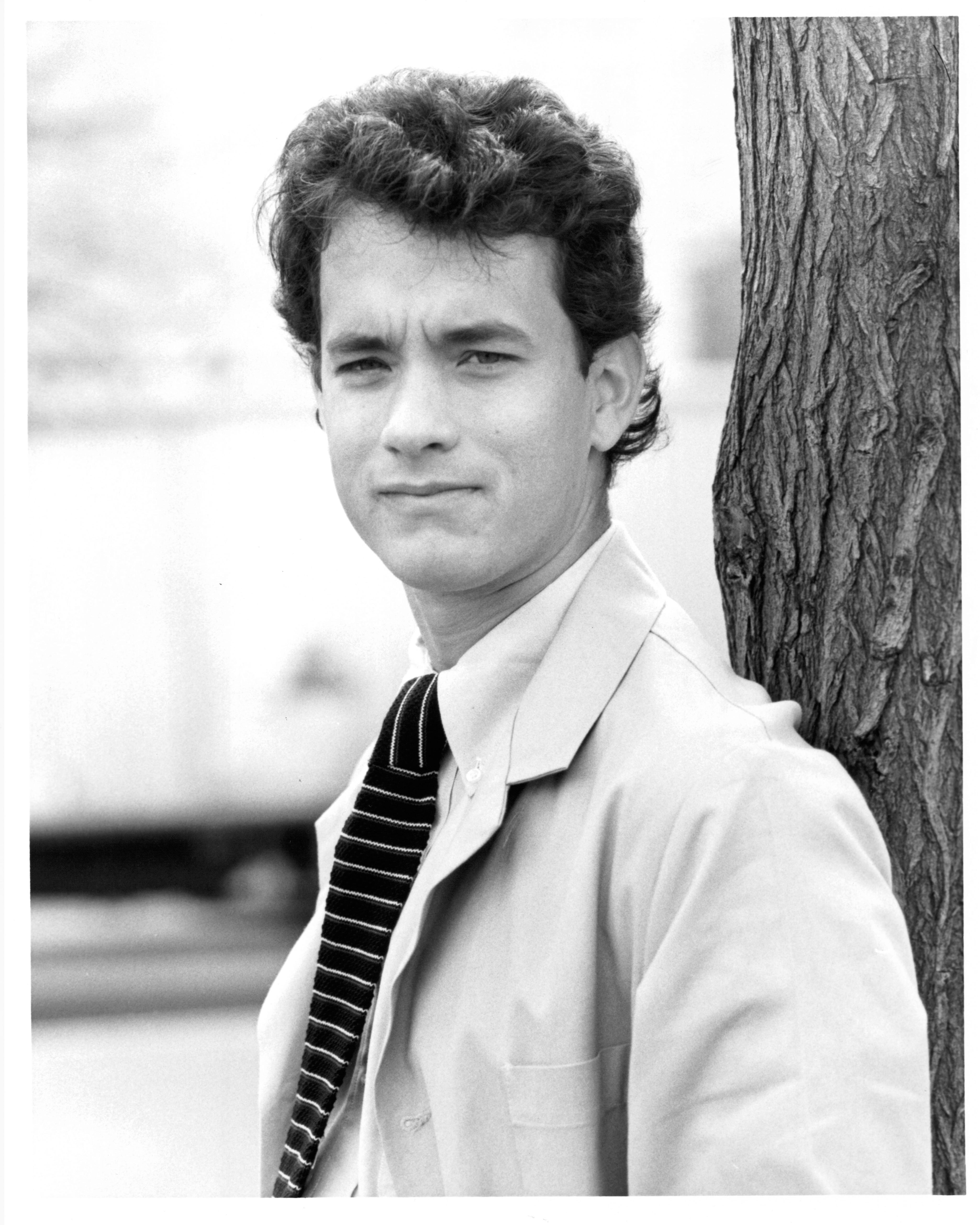 tom hanks looks at the camera as he leans his back against a tree trunk, he wears a collared shirt, tie and suit jacket