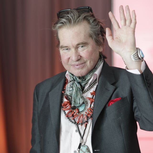 a man waves and wears a suit, a scarf, and necklaces