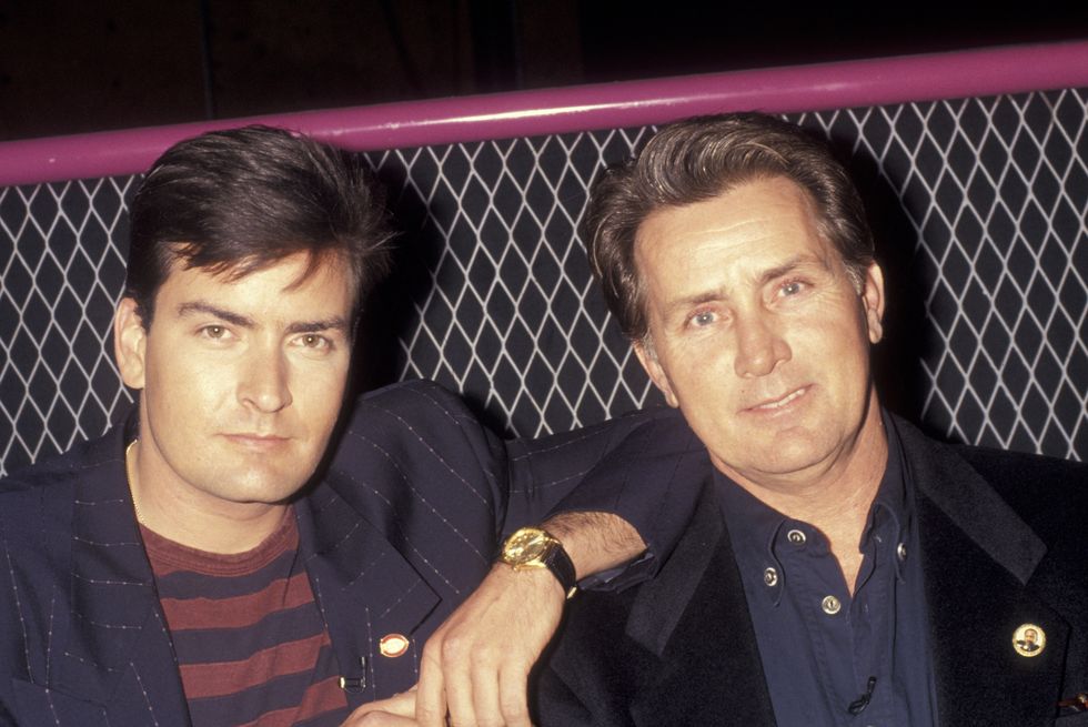 two men sitting next to each other, the man on the right rests an elbow on the other man's shoulder, both look at the camera