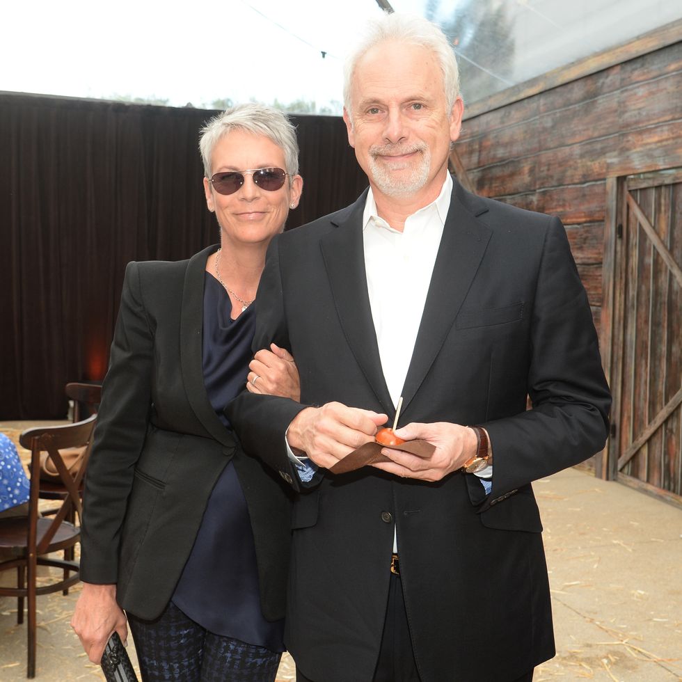 jamie lee curtis and christopher guest smile for a photo while standing together, they both wear black blazers and hold items in their hands