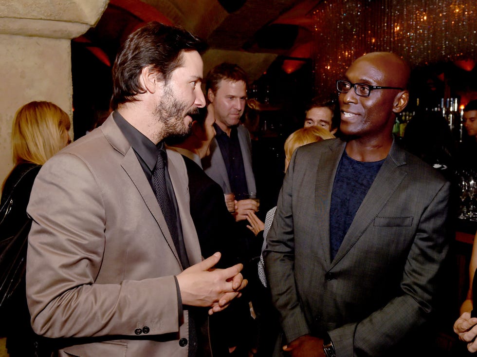 keanu reeves, wearing a tan suit, talks to lance reddick, wearing a black suit