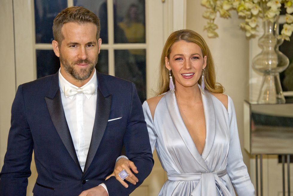 ryan reynolds and blake lively walk arm in arm inside a room, he wears a navy tuxedo jacket, white shirt and white bow tie, she wears a pale blue dress with a deep v and dangling earrings