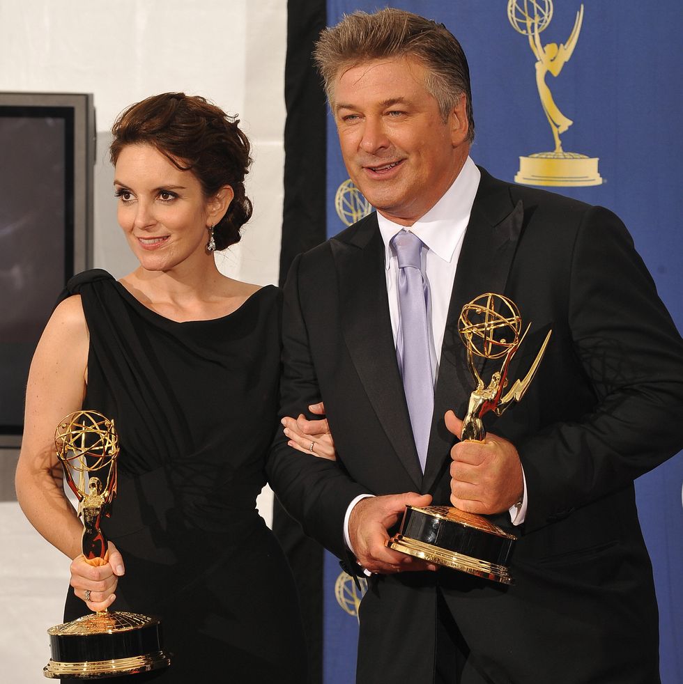 tina fey and alec baldwin each hold an emmy award and pose for photos while standing in front of a blue backdrop, she wears a black sleeveless dress, he wears a black suit with a white collared shirt and lavender tie