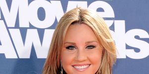 amanda bynes smiles at the camera in front of blue backdrop with white and neon green mtv movie awards logo, she has on heavy eye makeup, earrings, and a tan bandage dress, her long hair is curled
